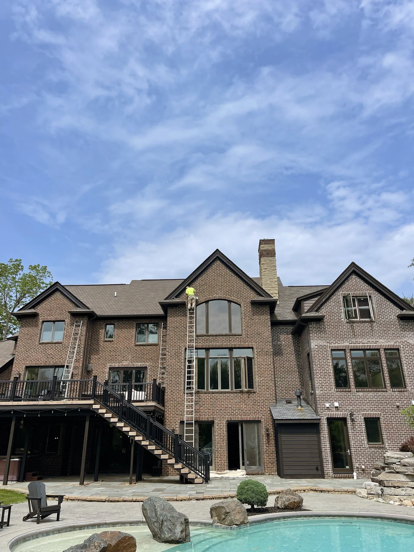 The rear of a large brick house with a deck and pool, two painters on tall ladders at the upper windows.