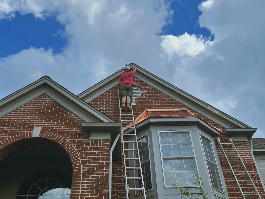 A painter on a ladder at the peak of a brick gable under a copper bay roof.
